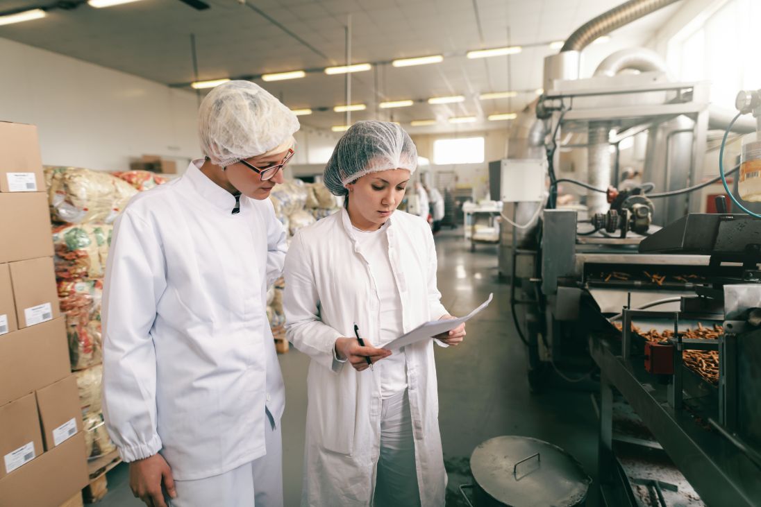 two quality professionals in white sterile uniforms checking quality of salt sticks while standing in food factory 1 2